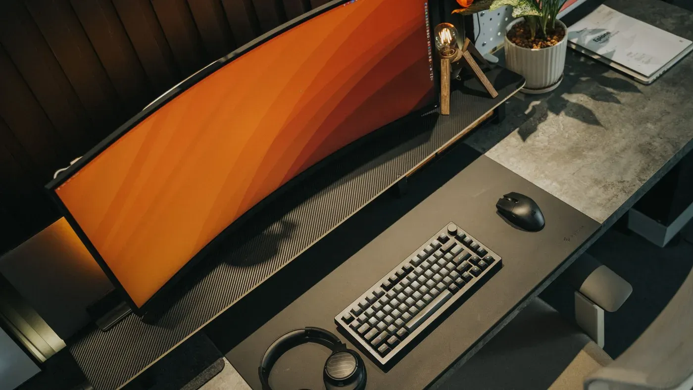 Gamingplats med böjd skärm, tangentbord, mus och hörlurar i neutrala toner.